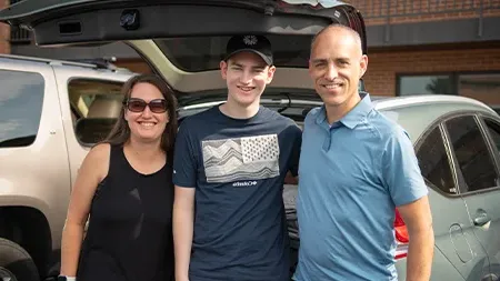 Student smiling with parents by car.